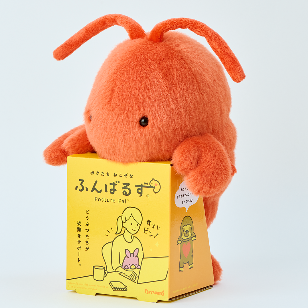 Image shows red/orange lobster Posture Pal in its yellow cardboard packaging which shows the product name in both English and Japanese as well as a cartoon image of a female sitting with a Posture Pal at her desk. The image is set on a white background. 