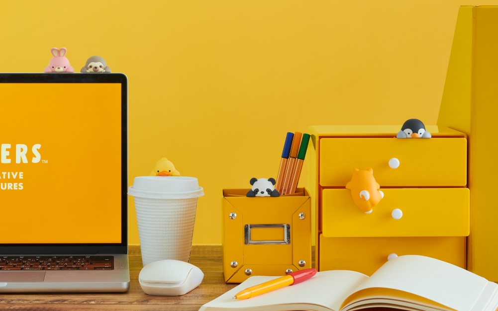 Image, set on bright yellow background, shows the new product lineup for Posture Pal Hippers. In the upper right corner of an open laptop sits the pink Rabbit and Sloth characters. To the right, atop a white cup sits the yellow Duck Hipper. To the right, the Panda Hipper sits in a yellow pen box, to the right, in the top drawer of a yellow three drawer set sits the Penguin Hipper and below, the Shiba Inu facing away. The image also shows a white computer mouse and open notebook with yellow and red pen. 
