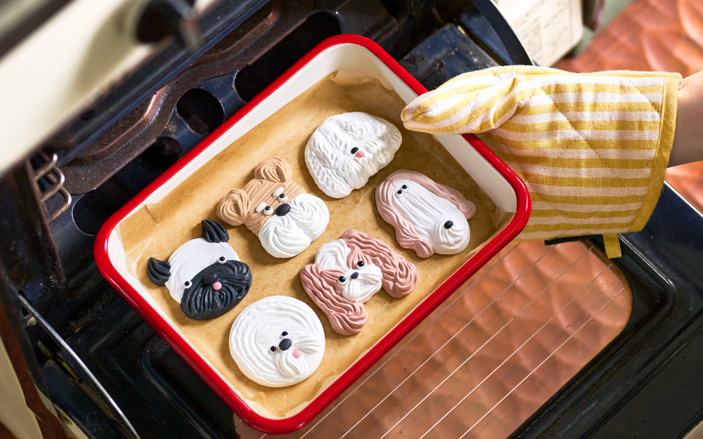 Image set in a kitchen, shows a person removing a tray of dog-shaped cookie stickers from an oven with a yellow and white striped glove. Left to right: black and white short nosed dog, tan and white short nosed dog, fluffy white dog, round fluffy white dog, tan and white long eared dog, and tan and white long face dog.