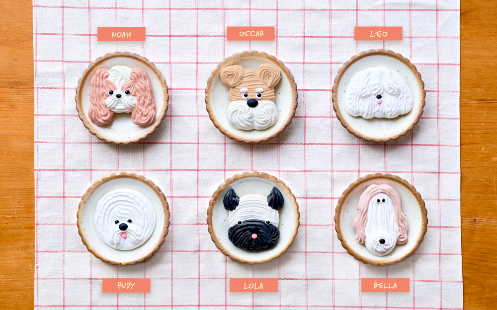 Image set on a wooden table with checkered tablecloth, six puppy themed cookie Patisstickers are each labeled by name. Left to right: "Noah" a peach and white long eared dog, "Oscar" a tan and white short nosed dog, "Leo" a fluffy white dog, "Rudy" a round short haired fluffy dog, "Lola" a black and white short nosed, pointy eared dog, and "Bella" a tan and white long snout dog
