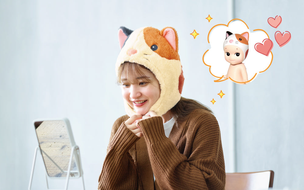 sonny angel costume series 2 product lifestyle image featuring a model wearing the CALICO CAT costume looking at themself in the mirror. illustrated speech bubble and hearts has a render of sonny angel calico cat minifigure