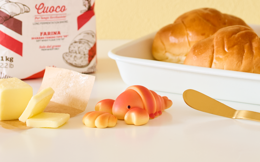 Toy croissant lobster next to a bowl of bread, cheese, and a box of flour on a white surface.