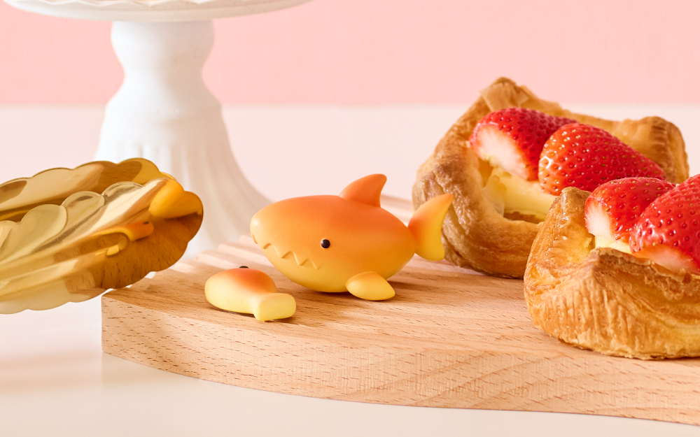 Small shark figurine next to strawberry pastries on a wooden board with a pink background