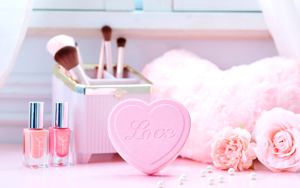 lifestyle image of rainbomb aurora heart floral scent on a pink table with nailpolish, rose, and makeup brush props. rainbomb says LOVE in the middle of the heart
