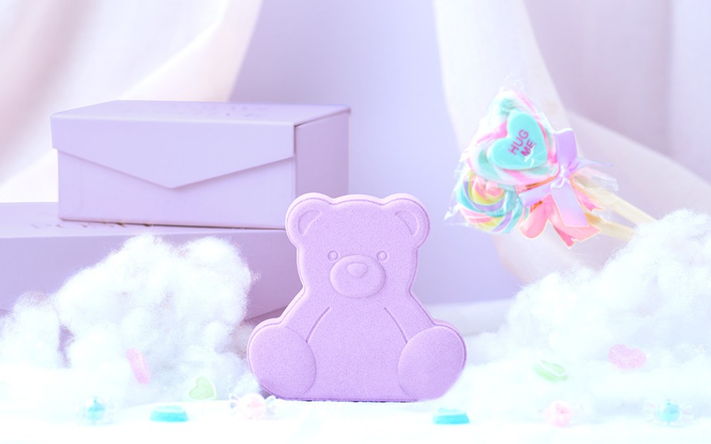 rainbomb aurora bear cotton candy product sitting on a bed of cotton balls with purple gift box and HUG ME conversation heart candy lollipop props
