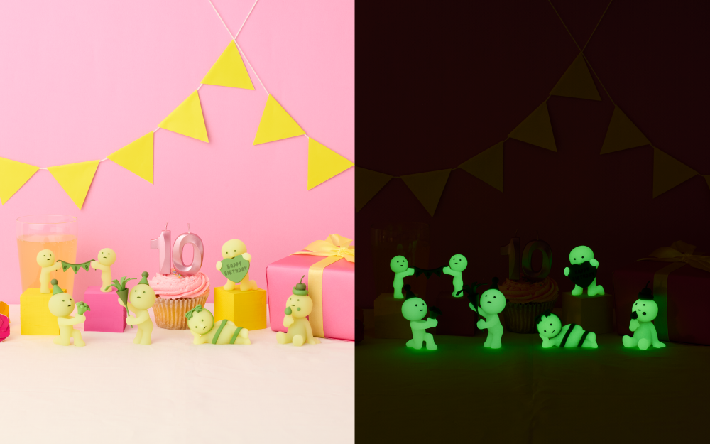 Split image set on pink background with yellow banner, pink frosted cupcake with candles and yellow drink, and pink gift. Image shows the new product line including SMISKIs holding a banner, SMISKI in party hat holding a bouquet, SMISKI holding a confetti popper with birthday hat, SMISKI holding heart with "Happy Birthday", and SMISKI lying down wrapped in ribbon beside SMISKI with a cherry on head eating frosting. Right side of image shows listed SMISKI glowing in the dark in the same positions and order. 