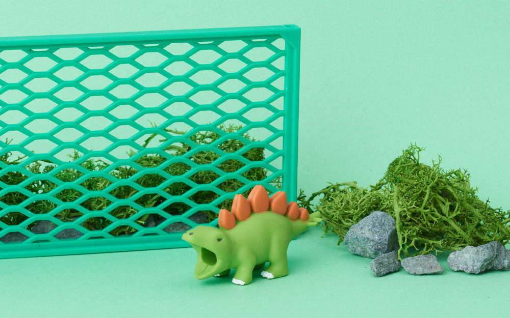 product lifestyle closesup of cable bite dino stegosaurus on a green background with fake moss, rock, and fence props