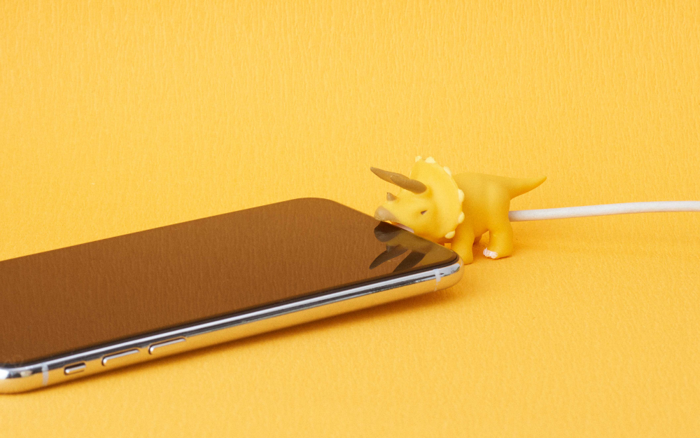 cable bite triceratops attached to a white cable and biting the bottom of an iphone. set against a yellow background