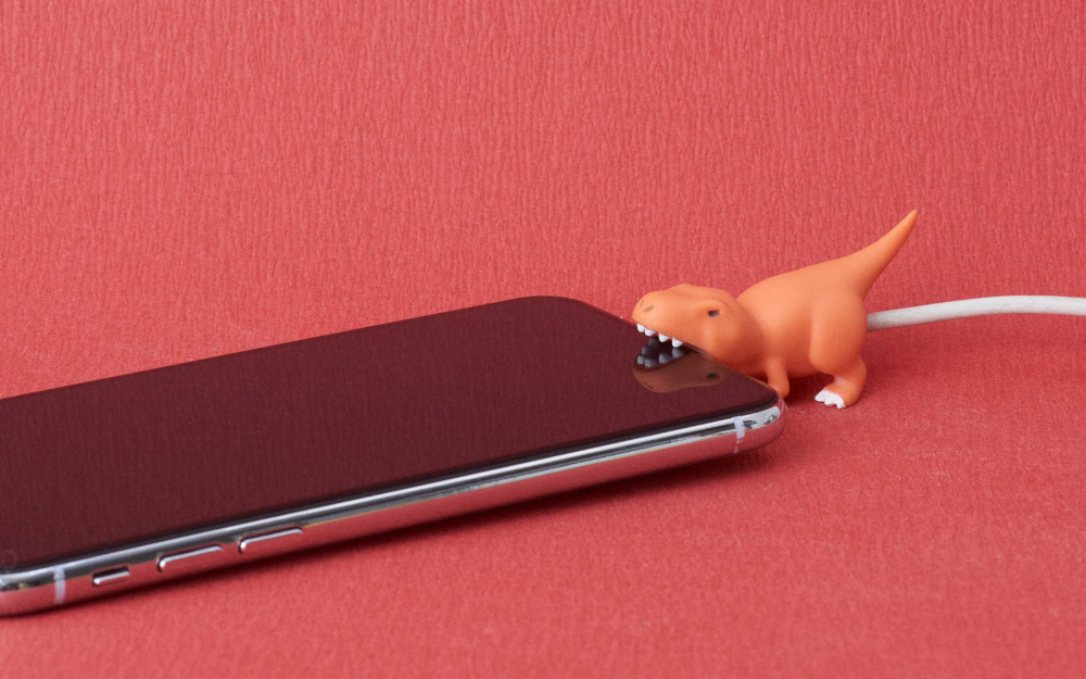 product closeup of cable bite dino trex on a white cable biting the bottom of a cell phone on a red background