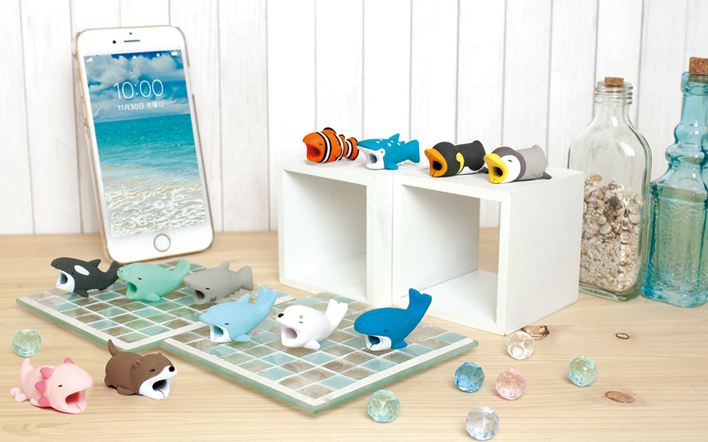 Image shows product variants for Cable Bite Volume 3. Set against a white wooden background, atop a wooden table, surrounded by colored gems, in front of an iPhone the variants are set. From left to right: Axolotl, Orca, Otter, Manatee, Shark, Dolphin, Seal, and Whale. On top of a white set of cube, from left to right: Clownfish, Whale Shark, Emperor Penguin and Penguin to the left of glass bottles filled with sand and liquid.