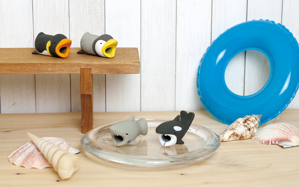 Image shows product variants for Cable Bite Vol 3. Set against a white wooden background, atop a wooden table, and shelf show the Emperor Penguin, and Penguin. In the center, the Shark and Orca sit on a glass plate in front of a blue floatation tube and seashells