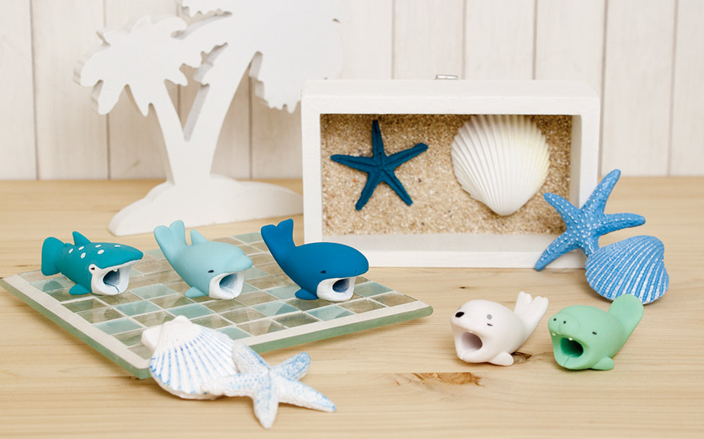 Image shows variants from product Cable Bite Vol. 3 staged against a white and tan background with ocean elements. A wooden palm tree, framed seashells, and table top seashells surround the Cable Bite variants. From left to right: Whale Shark, Dolphin, Whale, Deal and Manatee