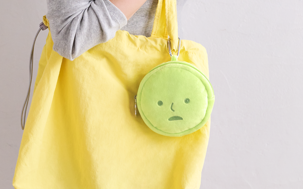 SMISKI plush zippered pouch hangs clipped to yellow cloth bag held by a figure in a grey shirt set against a grey wall