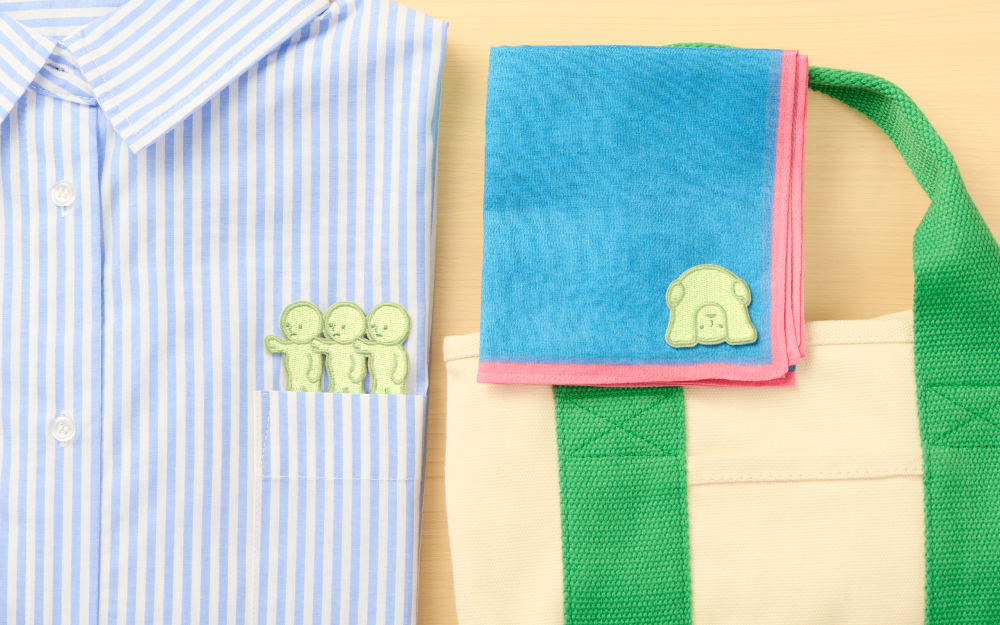 Left side of image, blue striped shirt with Little sticker variant above pocket, beside blue and pink folded towel with Peeking 2 sticker in bottom right corner atop green and tan canvas bag. Items set on wooden table background. 