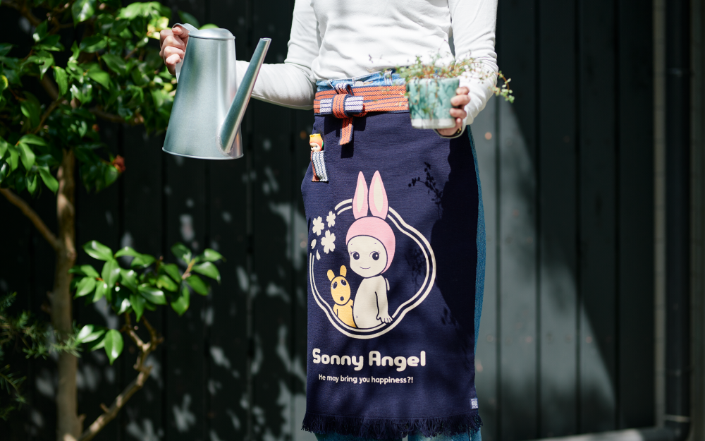Image shows a tree in the background beside a grey wooden fence. Figure in white shirt and denim jeans stands in the foreground holding a metal watering can and small potted plant. Figure is wearing the Maekake Apron with Tulip minifigure in apron's small red and white pocket. Apron's design: Rabbit Sonny Angel beside Robby encircled with cherry blossoms, and text: "Sonny Angel" "He may bring you happiness?!"