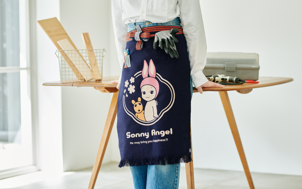 Image set in white room with large window. Center of the room is a wooden table with a metal basket holding pieces of wood, a tool case and construction tool. Figure stands in front of the table in a white shirt, denim jeans and "Maekake" apron. Work gloves are tucked into the red and white ties. Apron design: Rabbit Sonny Angel beside Robby Angel are encircled with cherry blossoms, and text: "Sonny Angel" "He may bring you happiness?!". A small red and white pocket holds the Rhino Sonny Angel minifigure. 