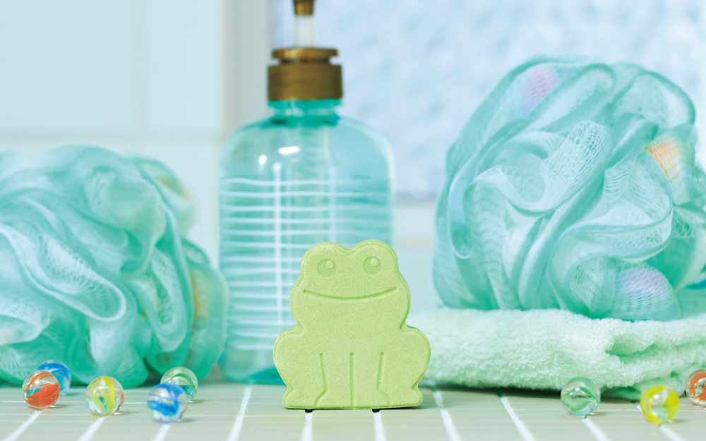 product closeup of rainbomb mini green frog on a green tile backdrop with loofah, marbles, soap bottle, and towel props