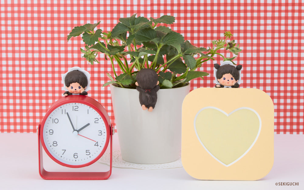 Monchhichi HIPPERS are set against a red checkered background on a white table. Figures from left to right include; baby boy in hood figure atop a red alarm clock, facing away figure on a potted plant, as well as the baby girl in hood HIPPER set on a yellow heart frame