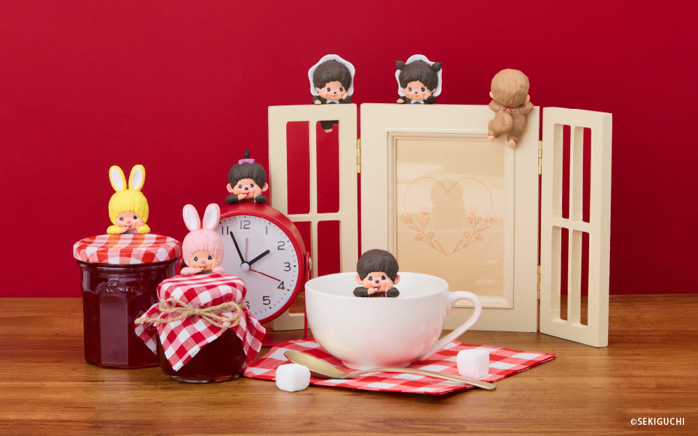 Monchhichi HIPPERS arranged against a red backdrop and wooden table. Yellow and pink bunny HIPPER set on jam jars beside the girl figure set on a red alarm clock, the boy figure sits in a white mug on a red checkered napkin. The back row includes three HIPPERS, baby boy in hood, baby girl in hood and tan HIPPER faces away, all set on a wooden frame