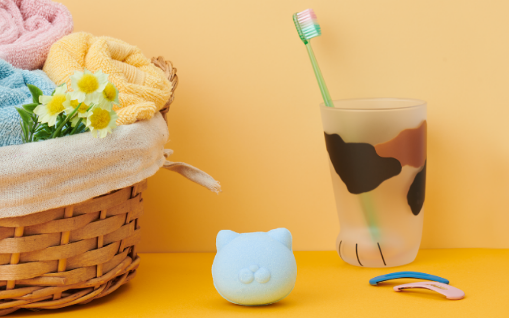 Product image for Nekomote Bath Bomb- Rosemary scent. Set against a light yellow background, atop a bright yellow counter, center image shows the  blue bath bomb product. To the left of the bath bomb sits a basket of towels, to the right a calico patterned cup with toothbrush, and hairclips