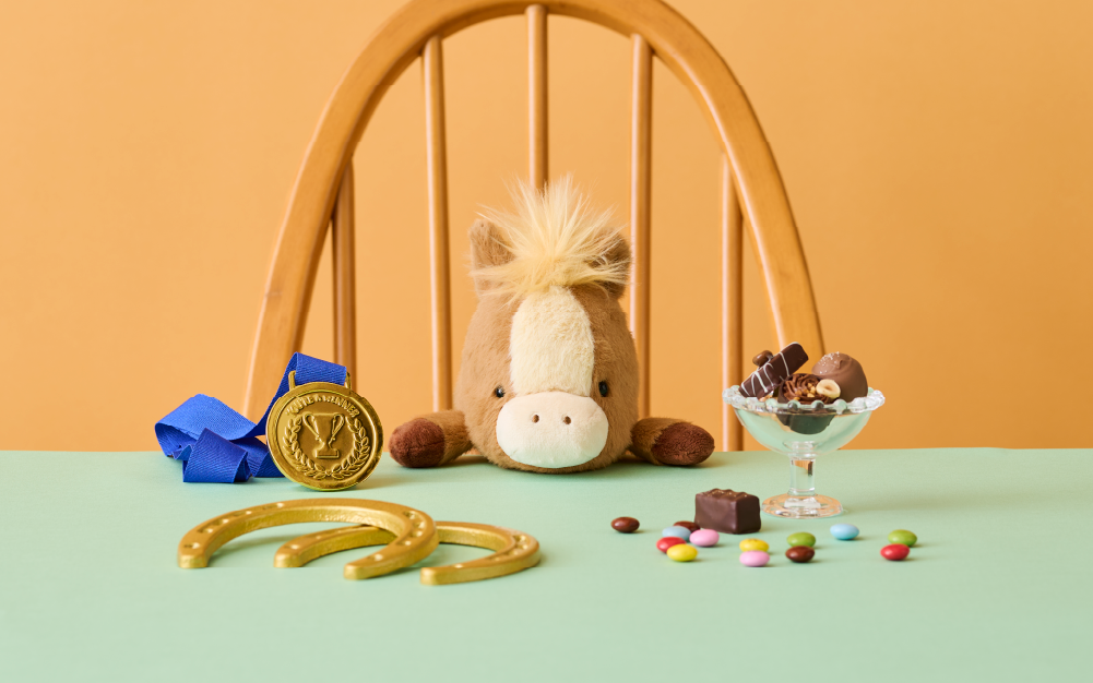 2026 Posture Pal Horse: Brown product shows the Posture Pal Horse on a wooden chair at a teal table top, against a yellow orange background. Atop the table, there is a blue ribbon gold medal, two gold horseshoes, and a variety of candies in a glass dish.