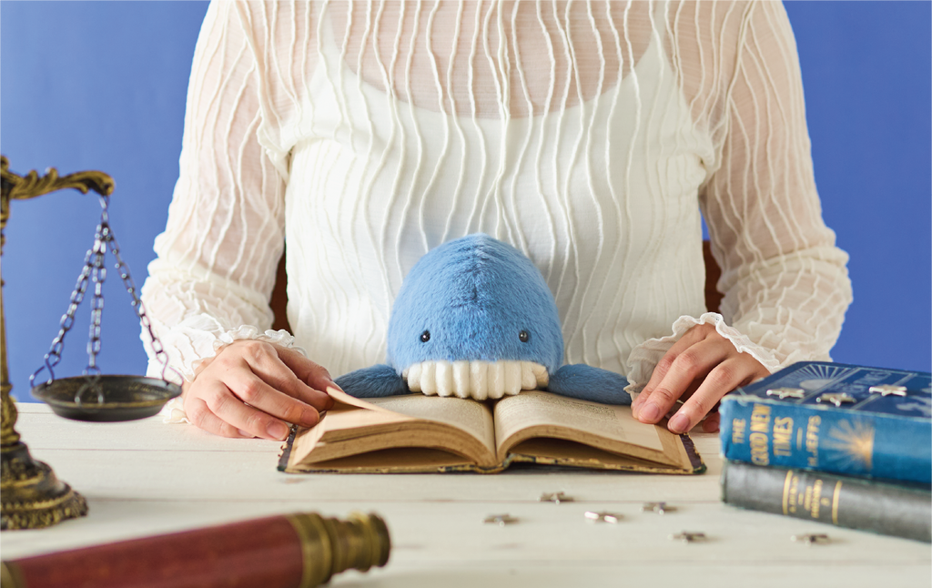 Image shows the upper body of a person in a textured white shirt sitting at a table with the blue whale Posture Pal. On the white table there are two closed books: one blue, one gray, one open book, a spyglass, silver star pieces, and partially shows a metal weigh scale. Image is set on a deep blue background.