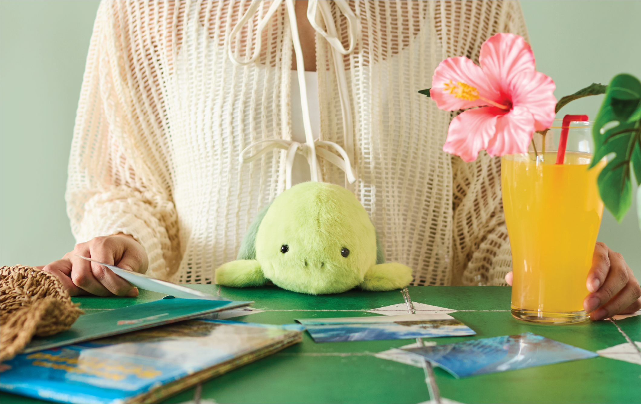 Image shows the upper body of a person in a knit top sitting at a green table. in the person's lap is the turtle Posture Pal, the person is looking at photos, there are photos and a folded map on the table, beside an orange tropical drink with a pink flower. the image is all set on a light green background