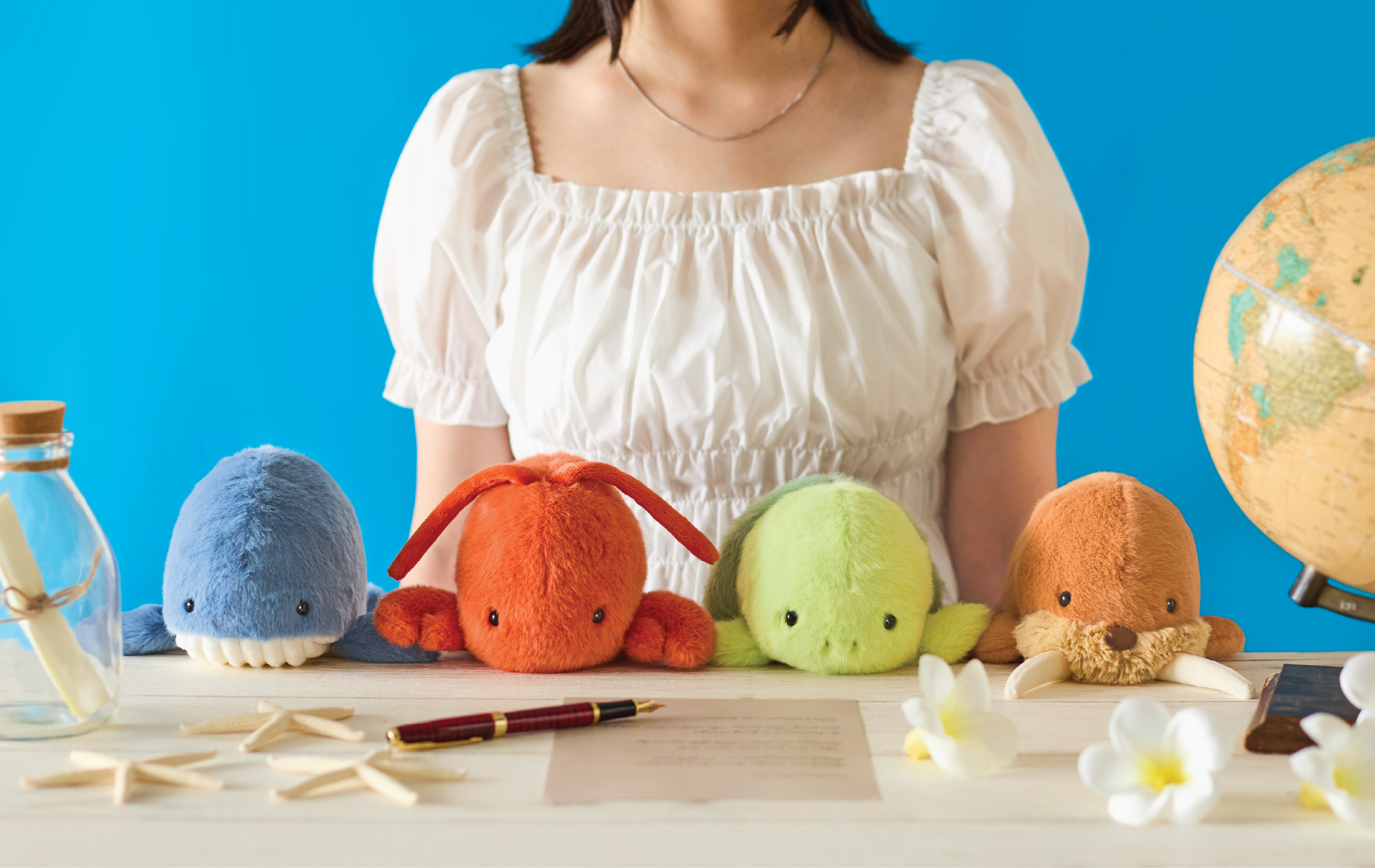 Image shows the upper body of a person in a white shirt sitting with colorful Posture Pals- left to right: blue whale, red/orange lobster, green turtle, brown walrus and a globe, flowers, starfish, a pen and message in a bottle sit on a table against a blue background