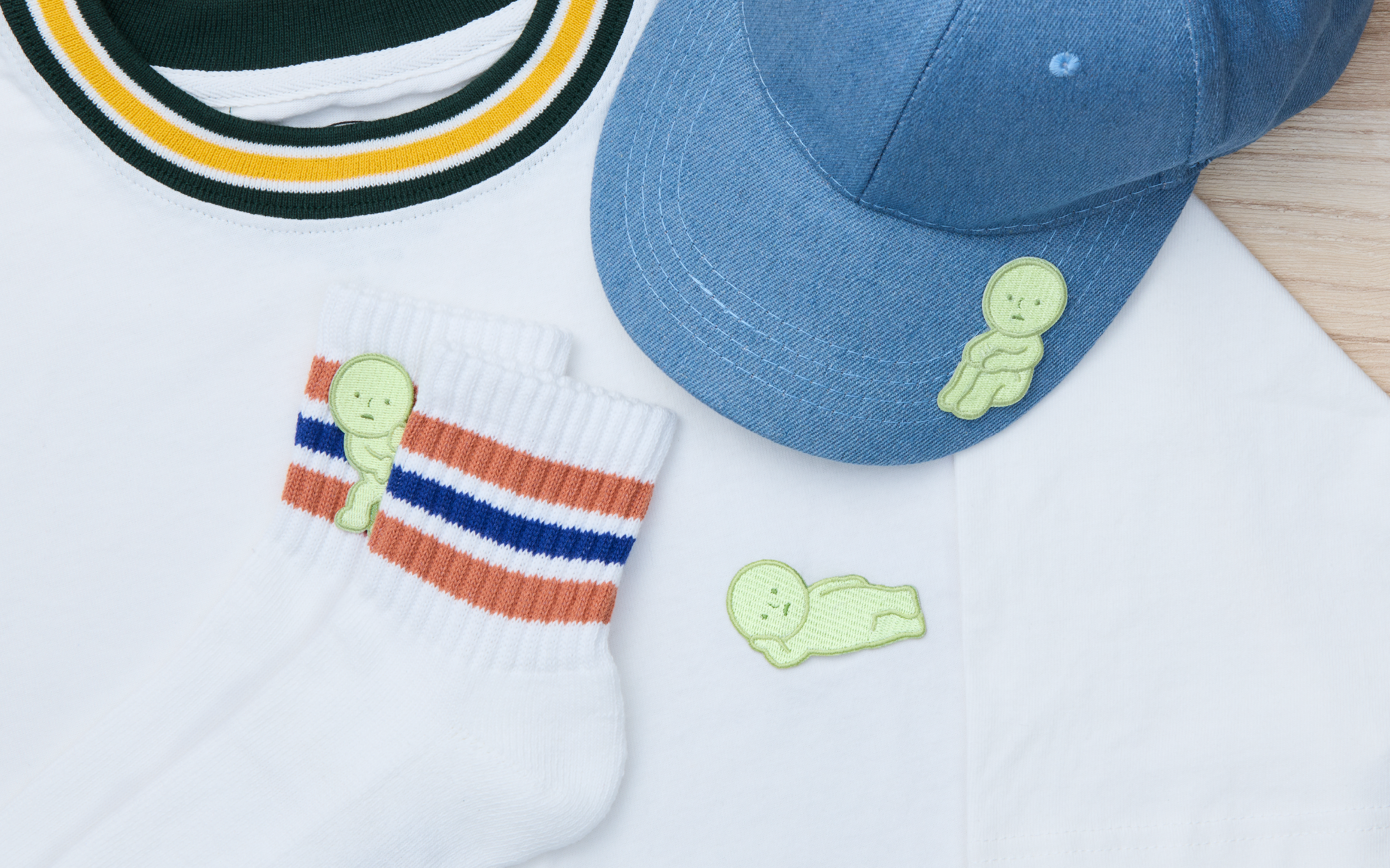 Image is set on a light brown wooden table. Atop the table is a white cotton shirt with a black and yellow collar. White cotton socks with red and blue striped cuffs show SMISKI "Peeking" Embroidery Sticker. On the white cotton shirt, SMISKI Embroidery Sticker "Lounging" is shown and above that is a blue denim baseball cap with the SMISKI Embroidery Sticker "Hugging Knees" on the right side of the brim. 