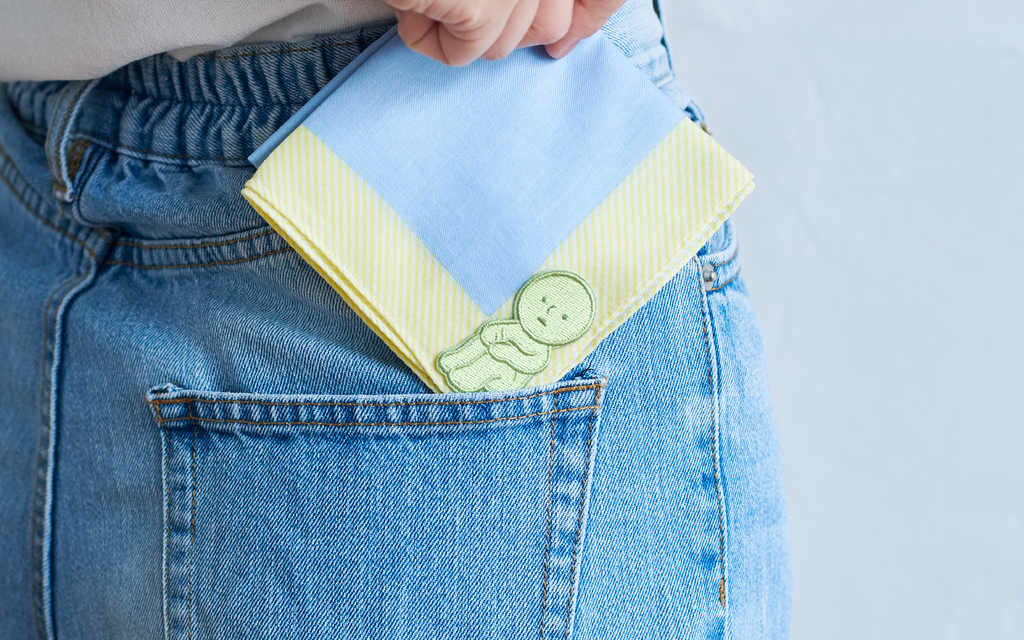 Image set on a light blue background shows the back of a person in a white shirt and blue denim jeans holding a light blue and yellow folded handkerchief. Adhered to the bottom right corner of the handkerchief is the SMISKI "Hugging Knees" Embroidery Sticker.
