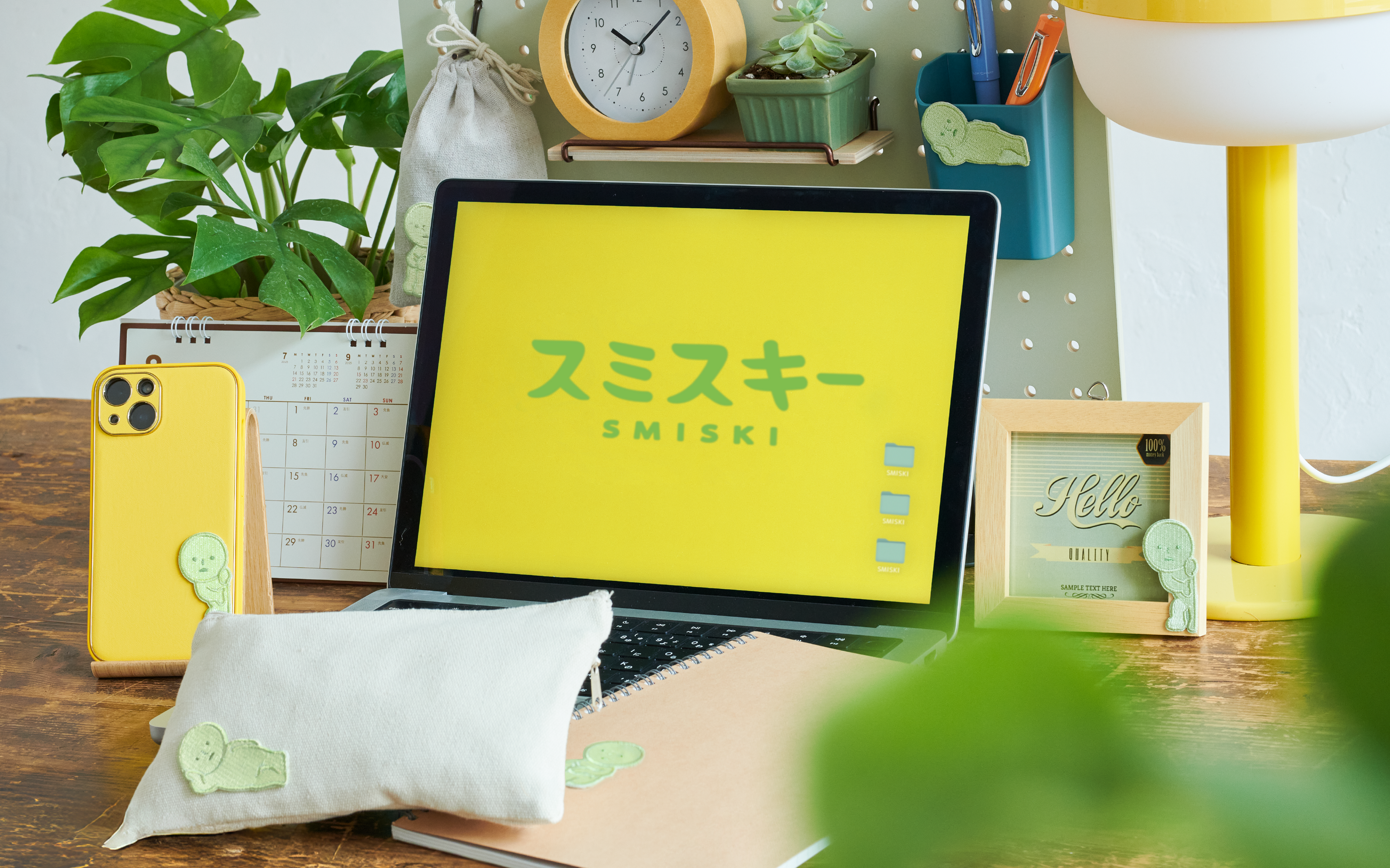 Image set on a wooden table with white walls. Atop the wooden table are objects such as potted plants, a yellow clock, pen holder with "Lounging" Embroidery Sticker and yellow lamp. From L to R: a yellow phone case with SMISKI Embroidery Sticker "Peeking", white canvas zipper pouch with SMISKI Embroidery Sticker "Lounging" on an open laptop with a yellow screen shows green SMISKI logo. Beside, a tan notebook with SMISKI Embroidery Sticker "Hugging Knees", and photo frame with "Peeking" Embroidery Sticker