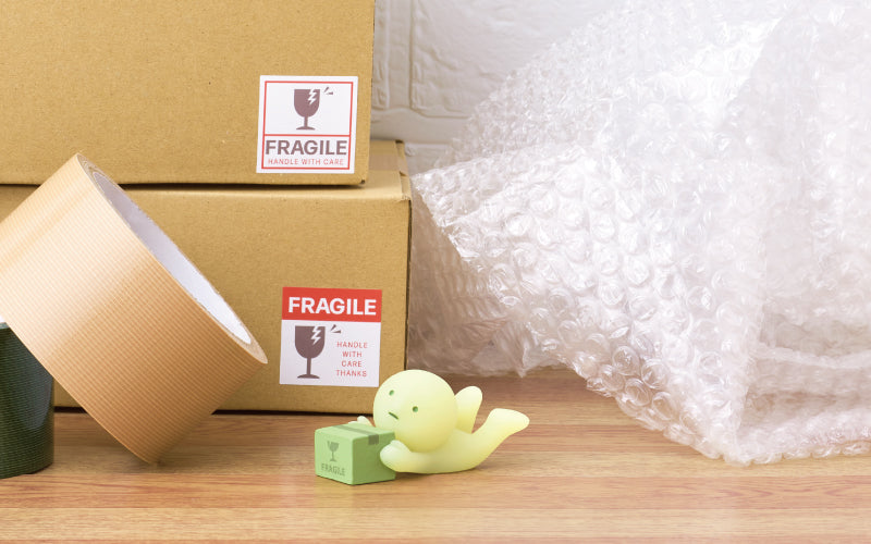 SMISKI Moving Series image set against a white brick wall, atop a wooden table. To the left of the SMISKI Falling Down minifigure are rolls of packing tape, and cardboard boxes, with bubble wrap to the right of SMISKI Falling Down