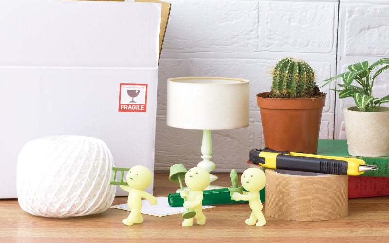 SMISKI Moving Series image set against a white brick wall, atop a wooden table shows moving boxes, white rope, a small lamp, packing tape, and potted plants. From left to right, SMISKI minifigures include: SMISKI Carrying Ladder, SMISKI Decorating, and Green Thumb SMISKI