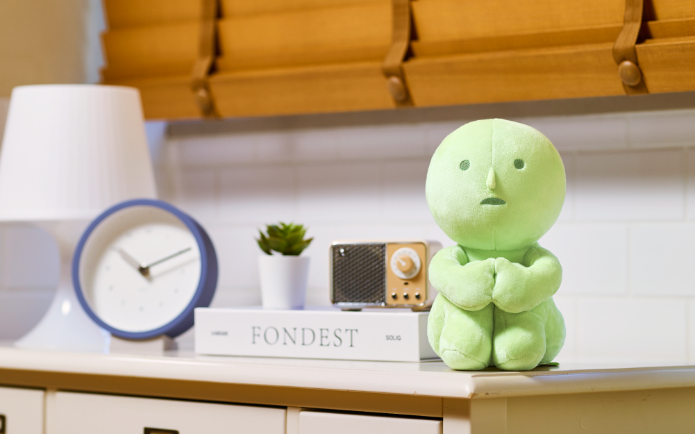 "Hugging Knees" SMISKI plush sits hugging knees to chest on countertop in front of a lamp, clock, book, small plant and radio. 