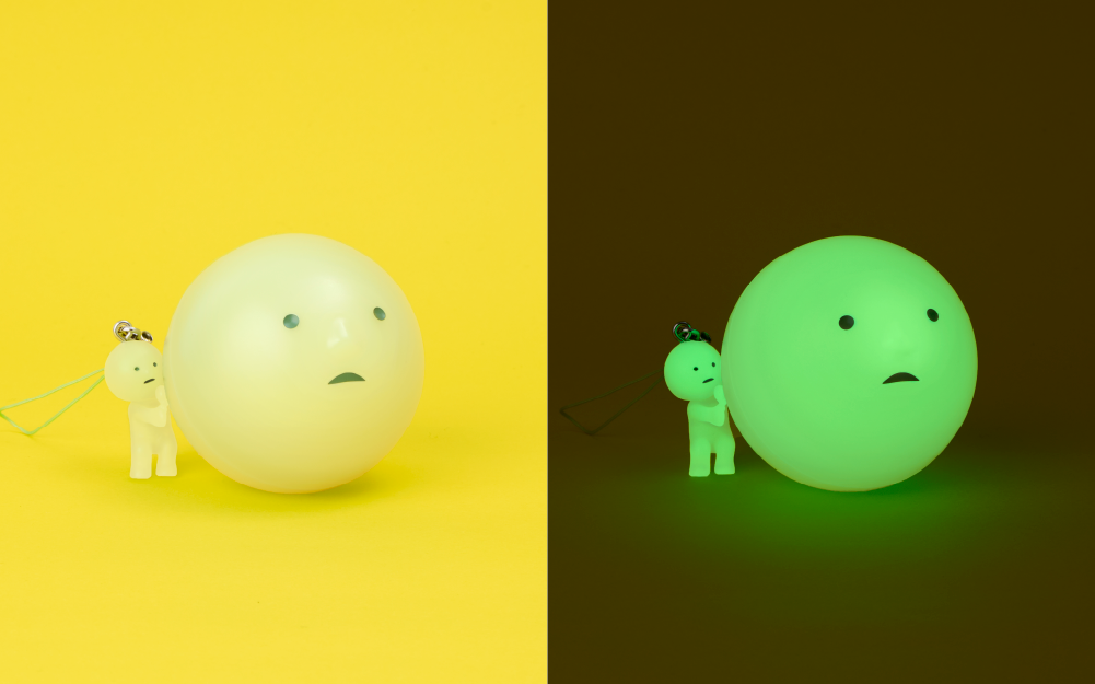 smiski strap accessory lifestyle image split down the middle. left: smiski gachapon capsule with a SMISKI peeking leaning against the capsule. both are on a yellow background. right: the same image in the dark with the smiski gachapon capsule and smiski peeking charm figure both glowing green in the dark.