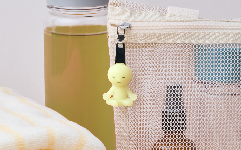 SMISKI Zipperbite "Lotus Pose" figure hangs from a white mesh zipper pouch, to the left is a glass jar of green tea, and a rolled up white and yellow striped towel. These items are set on a white background.