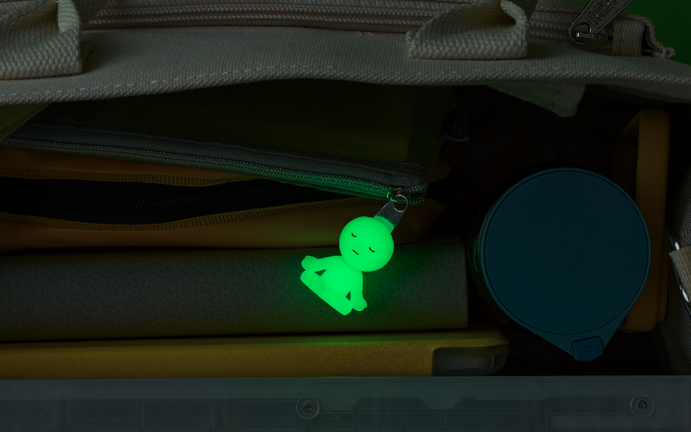 Glowing in the dark, "Lotus Pose" SMISKI Zipperbite hangs from a zipper pouch tucked into a white canvas bag, with books, and a drink bottle. 