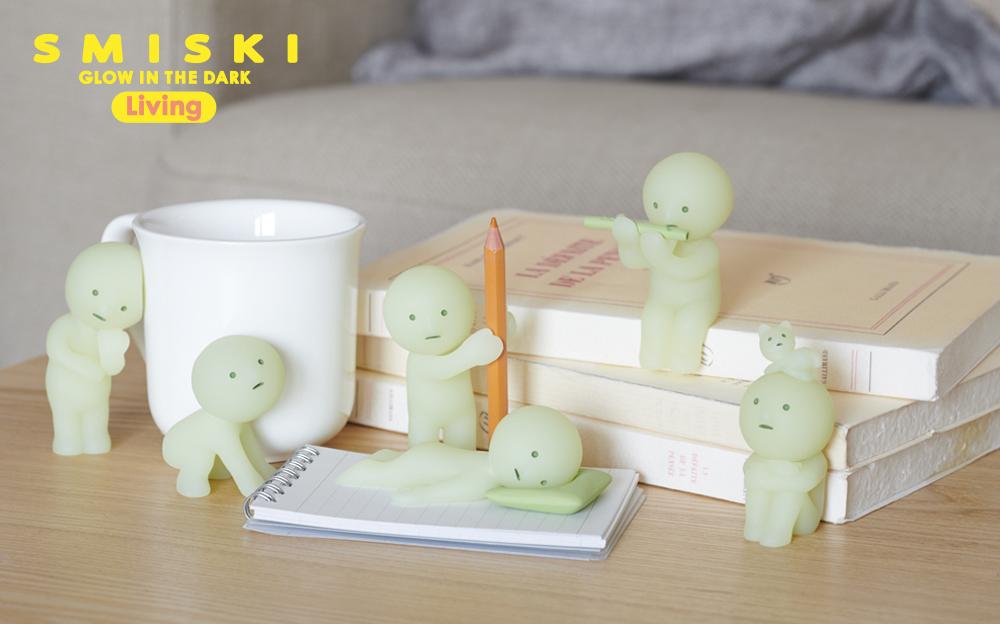 lifestyle image of smiski living full lineup on a desk backdrop with book, mug, and notebook props. text SMISKI GLOW IN THE DARK LIVING upper lefthand corner