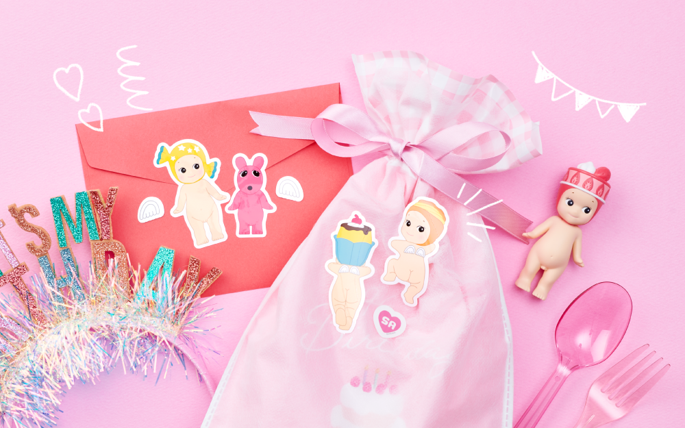 product lifestyle image of the sonny angel sticker pack series 2 stickers featuring stickers from the sweets pack placed on top of various party items like the back of an envelope and a gift bag. the overall theme of the image is PINK and there is a strawberry cake sonny angel mini figure in the center right of the image