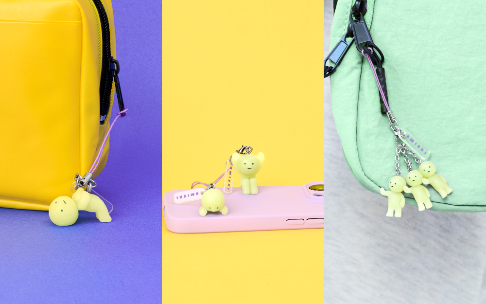 SMISKI Capsule Strap Vol 3 minifigures displayed in a image split three ways. Left: SMISKI doing a bridge connected to a yellow bag and black zipper. Middle: SMISKI peeking over the edge of a purple cell phone, beside SMISKI holding their hands up. Right: the little SMISKI trio hold their hands out, connected to a green bag and black zipper