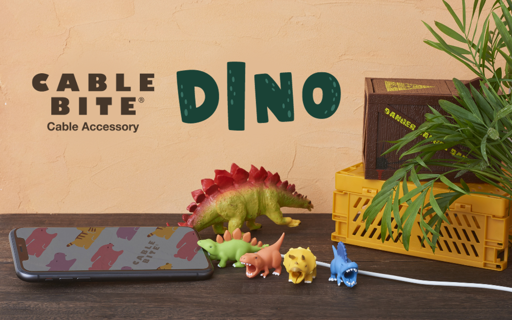 A series of dinosaur-shaped cable bites attached to a white cable, displayed on a table with various objects including a smartphone and a potted plant.