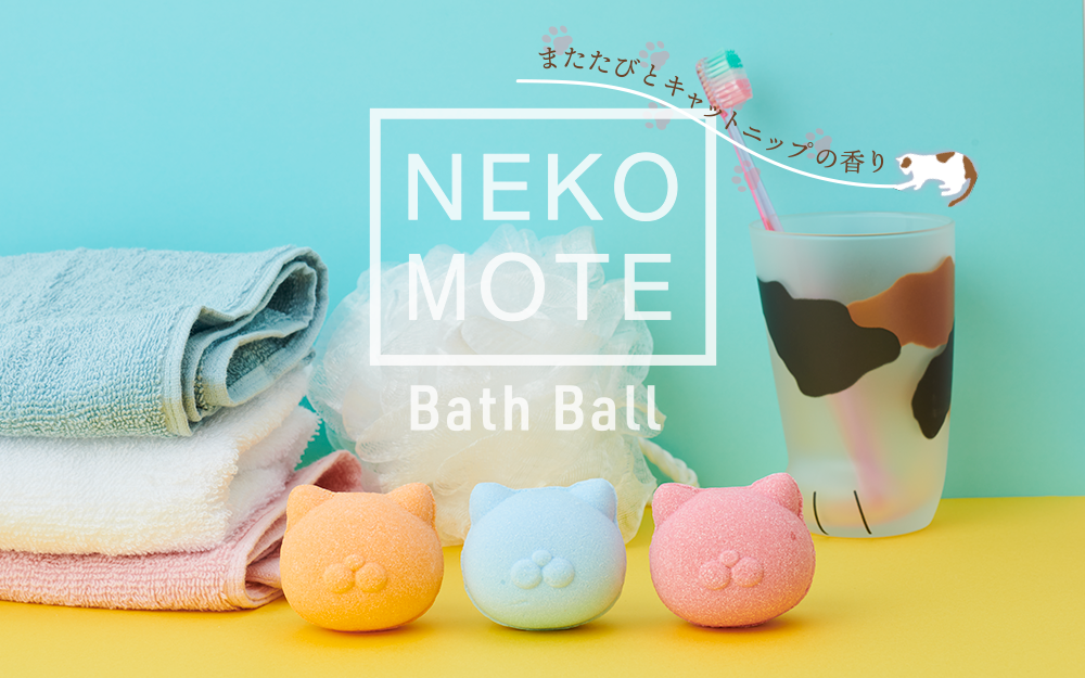 nekomote bath ball banner image featuring three nekomote bath balls (orange, blue, pink) removed from their packaging and displayed on a yellow surface in front of three towels, a loofah, and a cup shaped like a cat paw with a toothbrush inside. the product logo with the words NEKOMOTE BATH BALL and untranslated japanese text are overlaid across the image