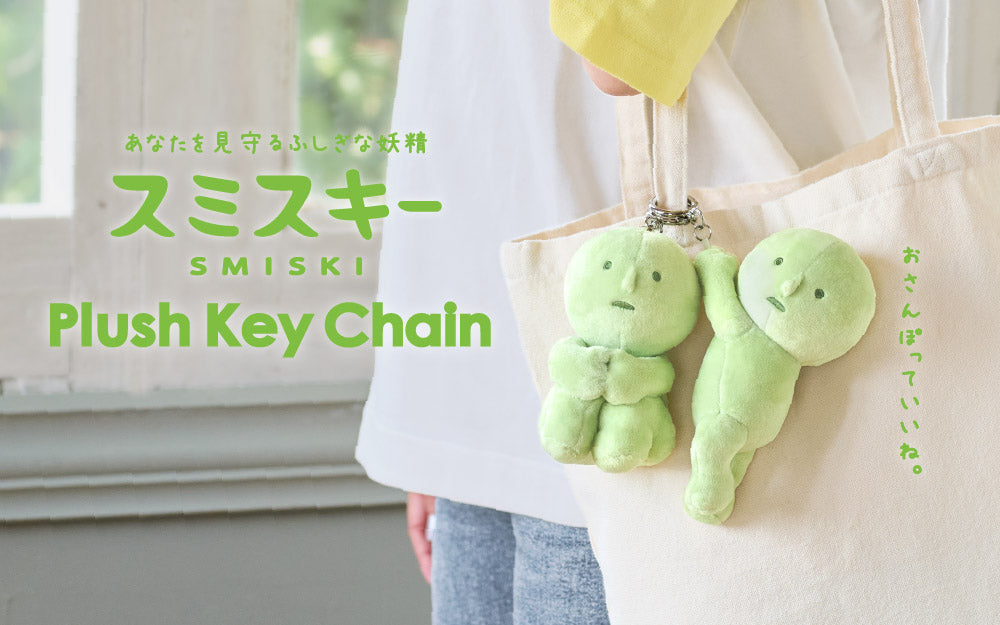 Image shows the two variations of product SMISKI Plush Key Chain written in both English and Japanese. (Right) depicts both variations of SMISKI clipped to a white canvas bag. SMISKI Hugging Knees to the left and SMISKI Hanging On to the right.