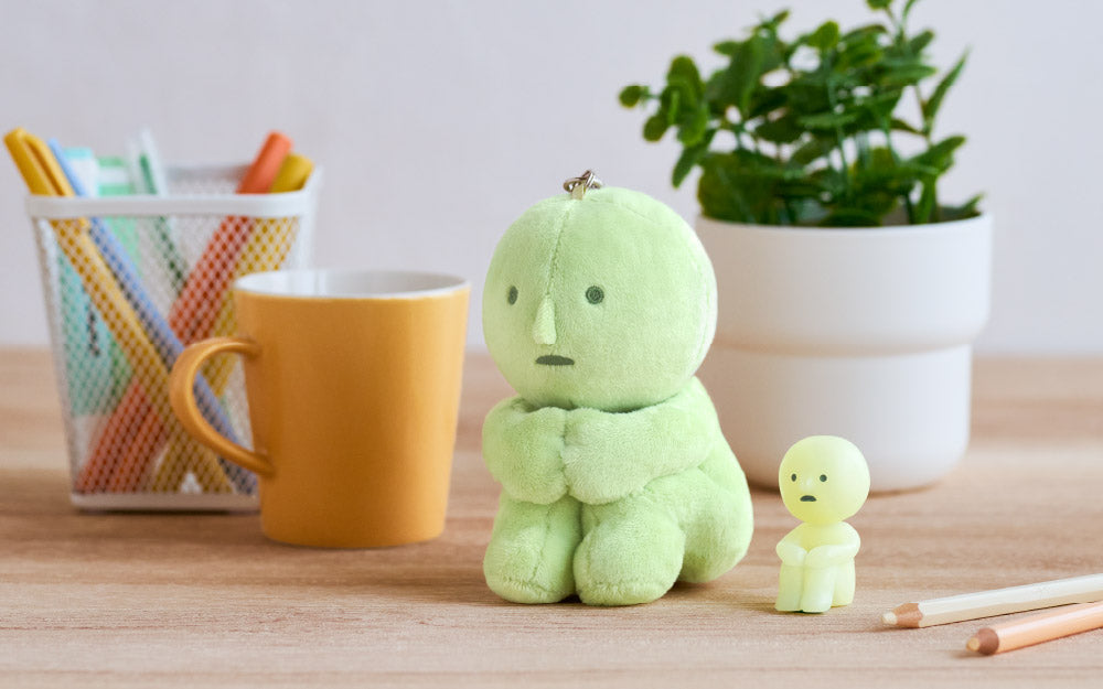 White background with a desk in the foreground. SMISKI Hugging Knees Plush Keychain (Middle) is in focus, with a SMISKI Hugging Knees minifigure (right) seated next to a white and a light pink colored pencil. Behind the two SMISKI items is a green leafy plan in a white pot. There is a white metal mesh pencil holder (left) out of focus with several pens of different colors. Directly between the pencil holder and the SMISKI Plush Keychain is a yellow mug.