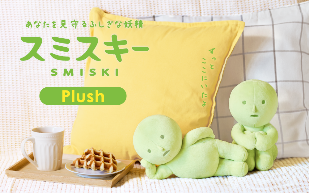 On a white bed background with yellow pillow, two green plush SMISKI figures are shown beside a tray with mug and waffles. Left: "Laying down" chubby plush lays on side with head propped up on hand. Right: "Hugging Knees" plush sits hugging knees to chest. 